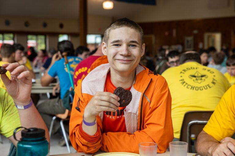 A smiling Scout sits in a camp dining hall holding a cookie, surrounded by other Scouts during a meal.