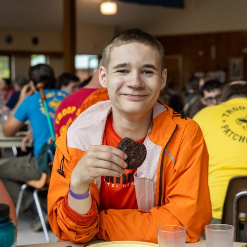 A smiling Scout sits in a camp dining hall holding a cookie, surrounded by other Scouts during a meal.
